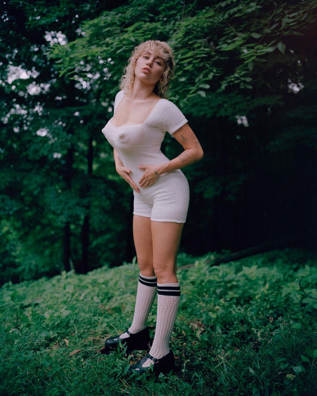 Young woman in a white romper and knee socks poses in a grassy, wooded outdoor setting, creating a blend of fashion and natural environment.