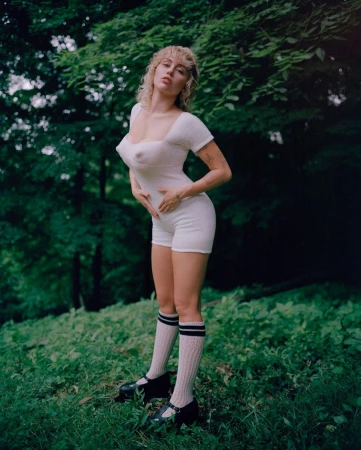 Young woman in a white romper and knee socks poses in a grassy, wooded outdoor setting, creating a blend of fashion and natural environment.