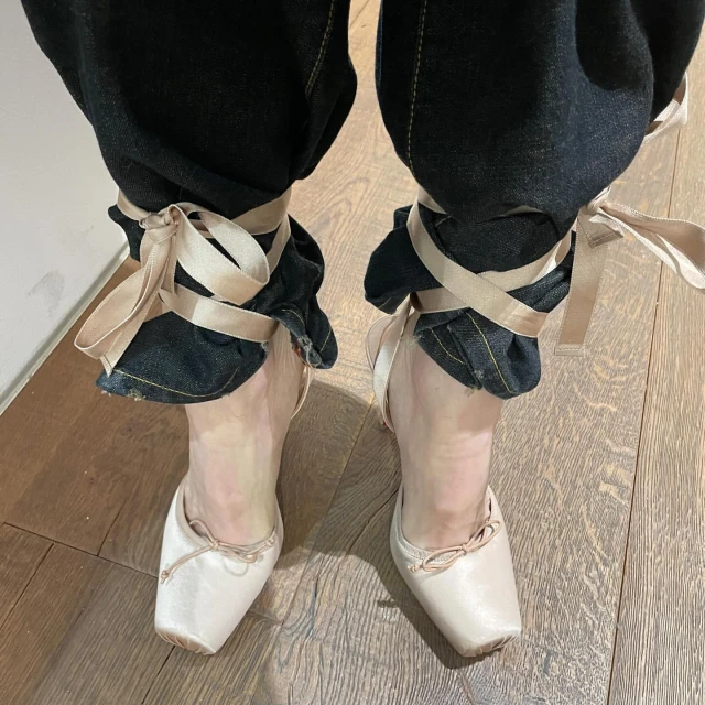 Close-up of human legs wearing dark denim jeans, satin ballet slippers, and satin ribbons tied around the ankles on a wood floor.