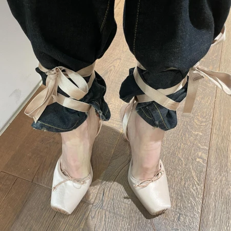 Close-up of human legs wearing dark denim jeans, satin ballet slippers, and satin ribbons tied around the ankles on a wood floor.