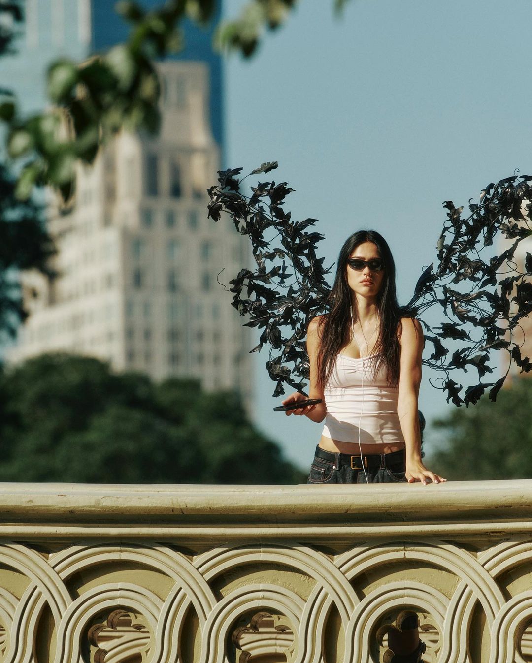 Young woman with decorative wings stands in an urban park with a building in the background on a sunny day, holding a cell phone.