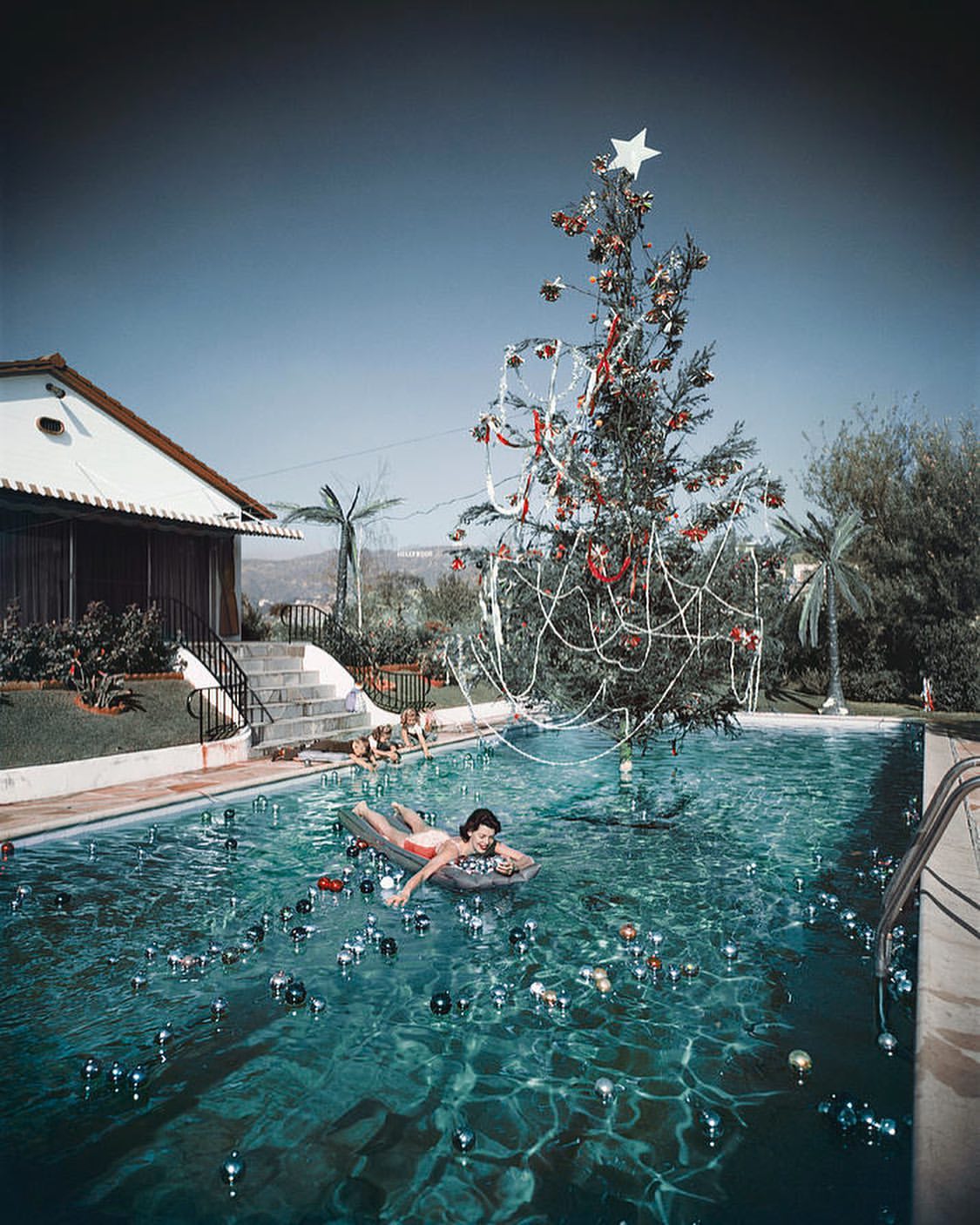 Woman floats on raft in swimming pool with Christmas tree, ornaments, and hillside homes in the background on a sunny day.