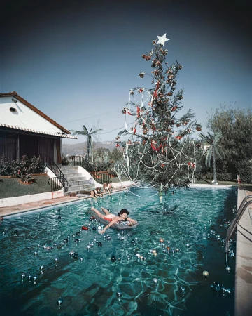 Woman floats on raft in swimming pool with Christmas tree, ornaments, and hillside homes in the background on a sunny day.