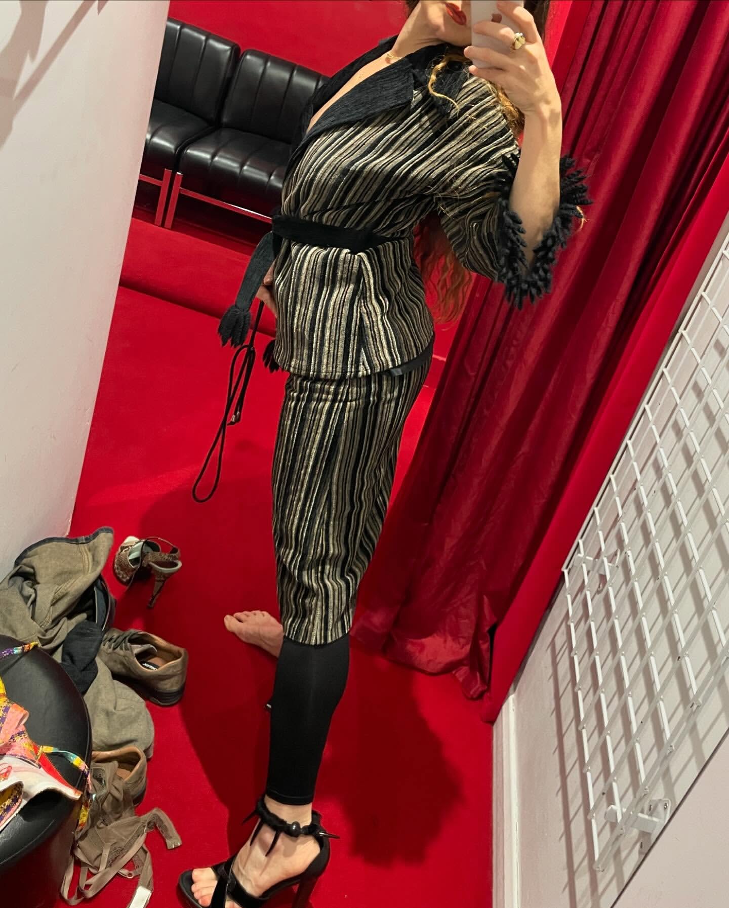 Adult female models a striped jacket and cropped pants with black leggings and high-heeled sandals indoors on a red floor.