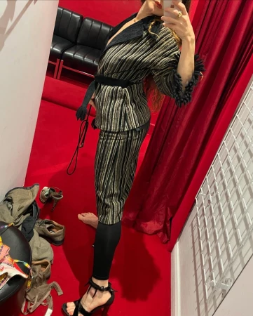 Adult female models a striped jacket and cropped pants with black leggings and high-heeled sandals indoors on a red floor.