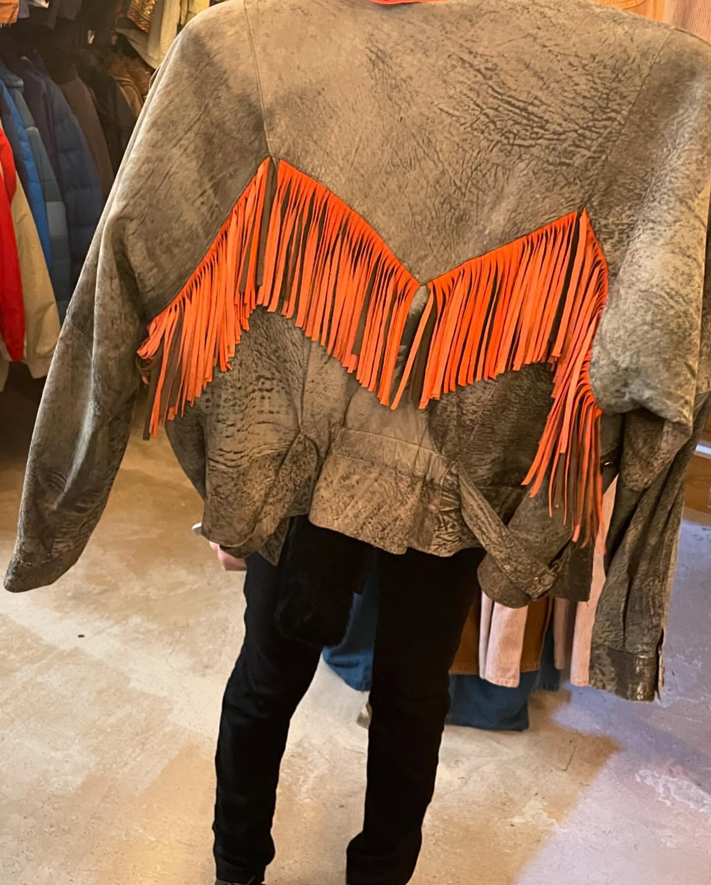 Grey suede jacket with orange fringe hanging in store, showcasing a unique fashion piece with a casual and vintage style.