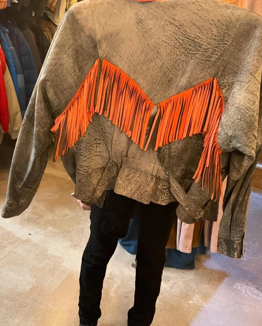 Grey suede jacket with orange fringe hanging in store, showcasing a unique fashion piece with a casual and vintage style.