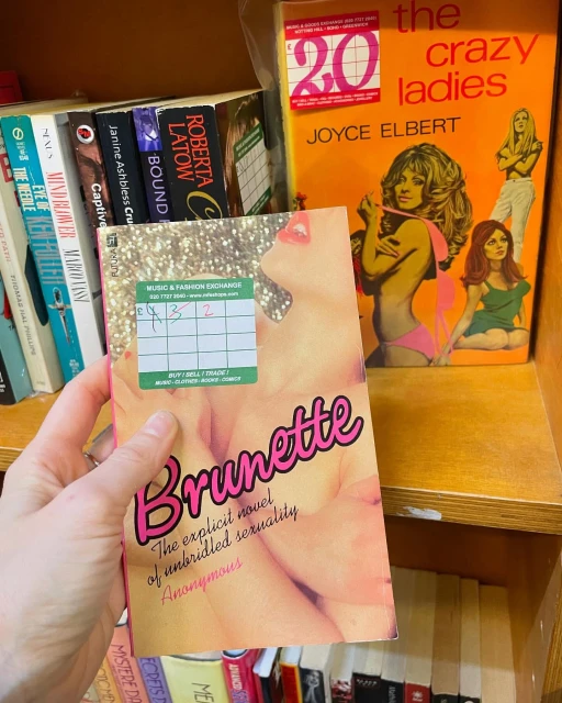 Close-up of a hand holding the book 'Brunette' in front of a shelf of books at Music & Fashion Exchange bookstore.