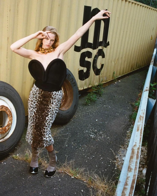 Fashion shot of a slender woman in a black bustier, leopard skirt, and gold necklace, posing near a shipping container and tires outdoors.