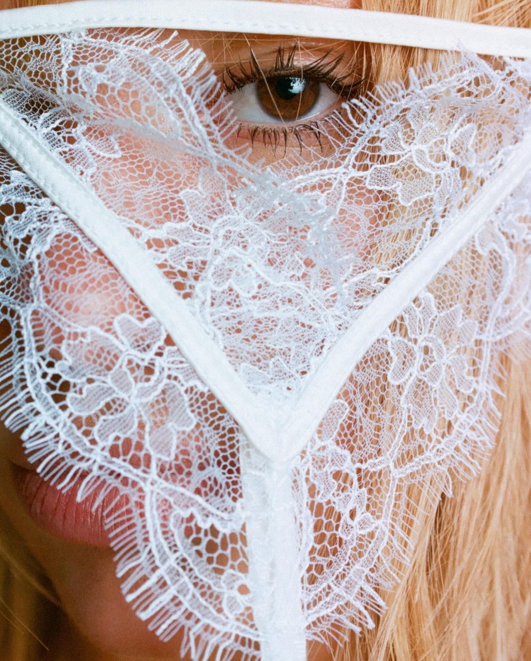 Close-up studio shot of a blonde woman wearing a white lace mask, emphasizing beauty and fashion details with soft lighting.