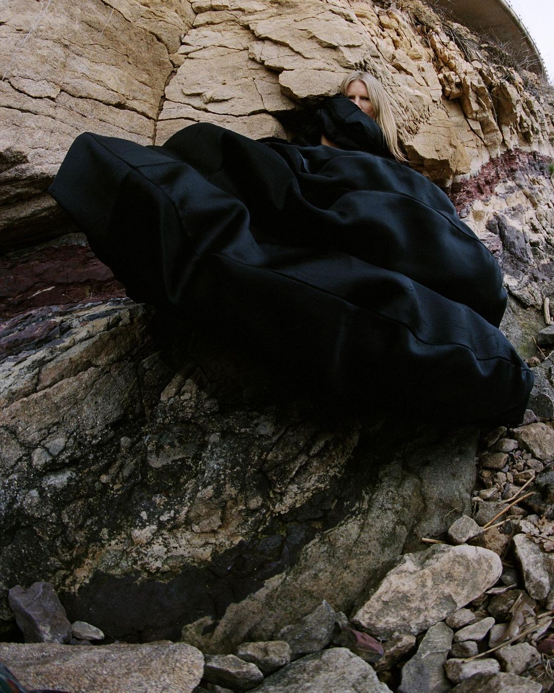 Model wearing a voluminous black garment partially hidden against a rocky stone cliff face