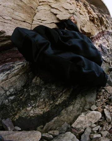 Model wearing a voluminous black garment partially hidden against a rocky stone cliff face