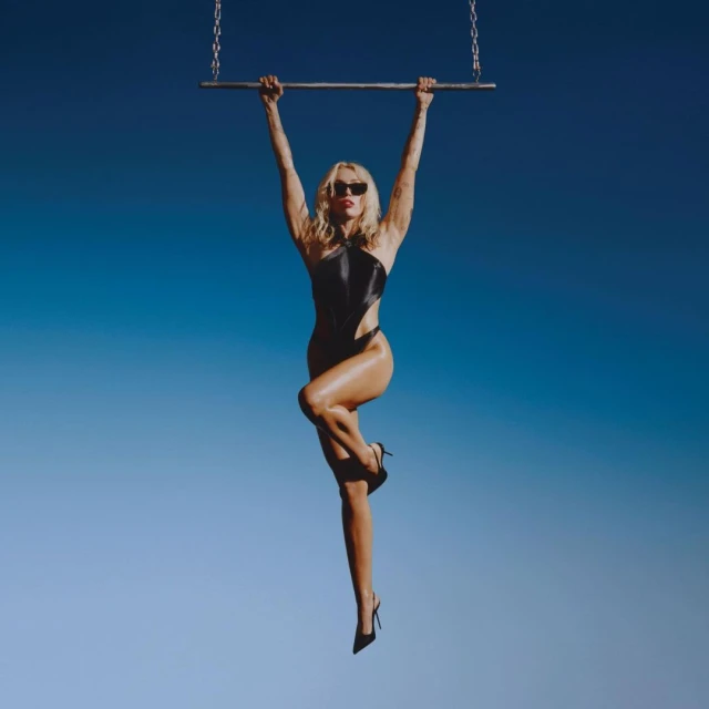 A woman with blonde hair and sunglasses hangs from a metal bar wearing a black bodysuit and high heels against a blue sky backdrop.