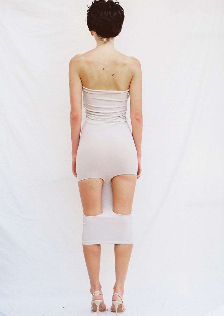 Rear view of a short-haired woman in a strapless beige dress and heels standing against a white seamless background.