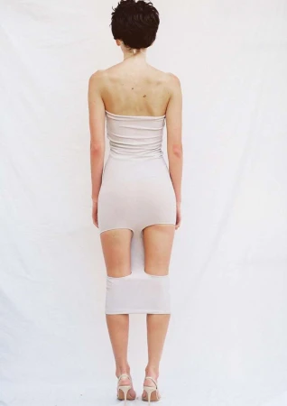 Rear view of a short-haired woman in a strapless beige dress and heels standing against a white seamless background.