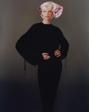 Model in a black sweater wearing a patterned pink head covering against a neutral grey background