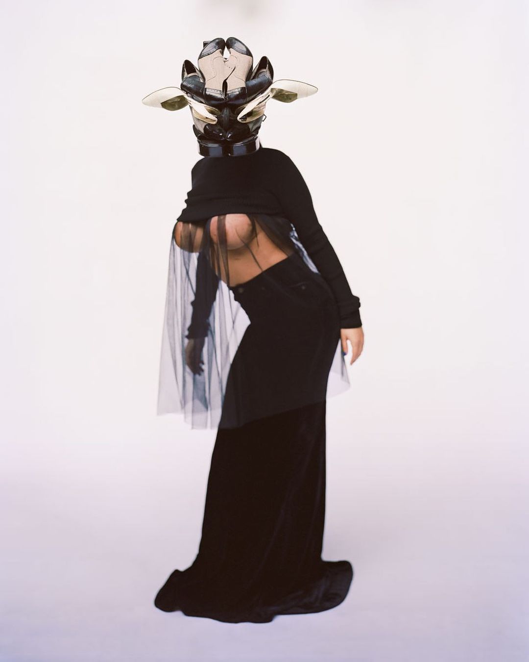 A woman in a conceptual fashion pose, wearing an elaborate mask, black top, black skirt, and sheer black cape against a white background.