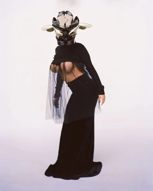 A woman in a conceptual fashion pose, wearing an elaborate mask, black top, black skirt, and sheer black cape against a white background.