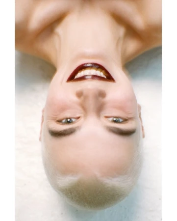 Upside-down close-up portrait of a woman with short blonde hair and dark glossy lipstick smiling against a white seamless background