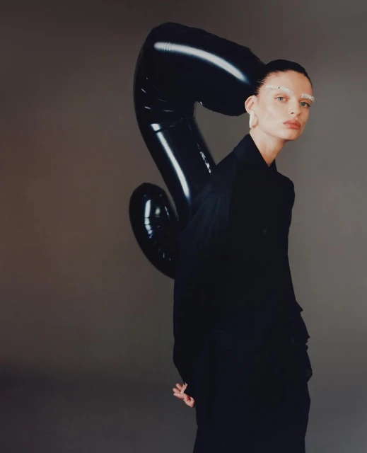 A woman with bleached eyebrows posing in front of a gray background with a black balloon next to her in a studio setting.