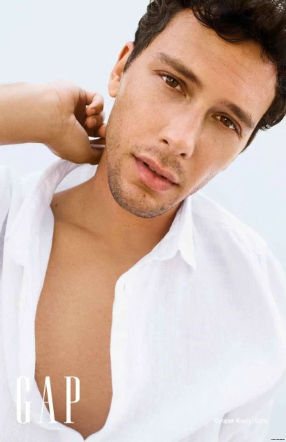 Close-up portrait of a handsome man with brown hair, wearing an unbuttoned white linen shirt, posing against a bright, soft background.