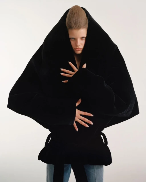 Fashion photo of a woman wearing a large, oversized black jacket with light-wash jeans in a white studio setting, looking at the camera.