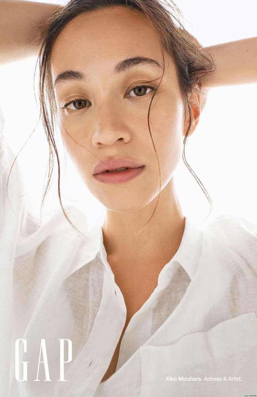 Close-up portrait of a young adult woman with wet hair pulled back, wearing an unbuttoned white shirt against a bright white background.