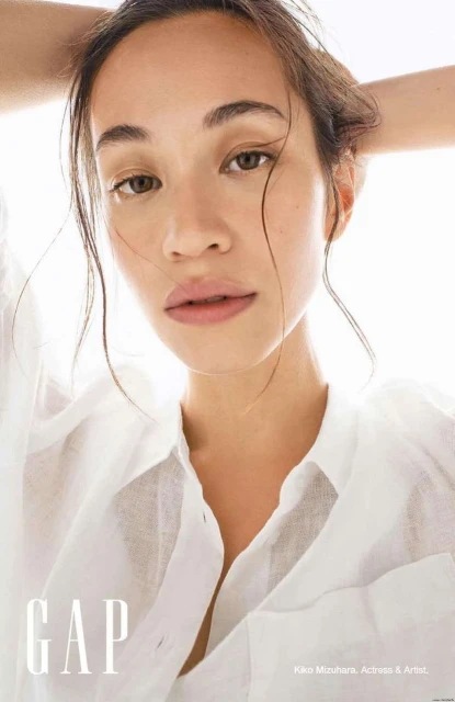 Close-up portrait of a young adult woman with wet hair pulled back, wearing an unbuttoned white shirt against a bright white background.