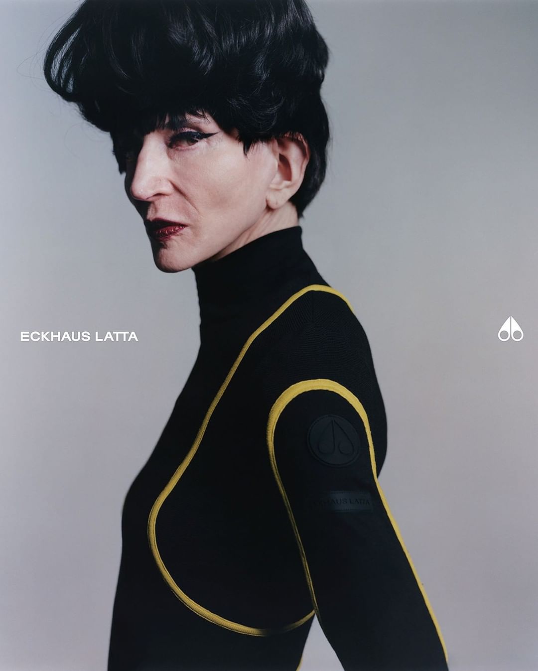 Headshot of an adult woman wearing black turtleneck top with yellow piping, with short dark hair and red lipstick in a studio setting.