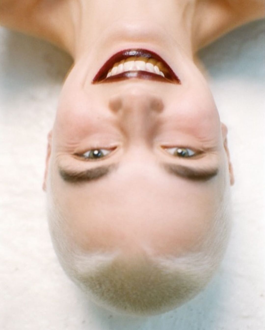 Close-up portrait of a woman with a shaved head, dark lipstick, and a slight smile, photographed from an upside-down perspective.