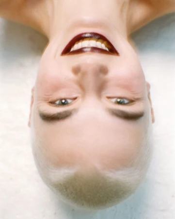 Close-up portrait of a woman with a shaved head, dark lipstick, and a slight smile, photographed from an upside-down perspective.