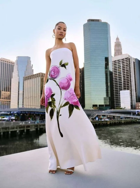 Woman in strapless dress with floral design stands in front of cityscape. Modern fashion shoot with an urban background.