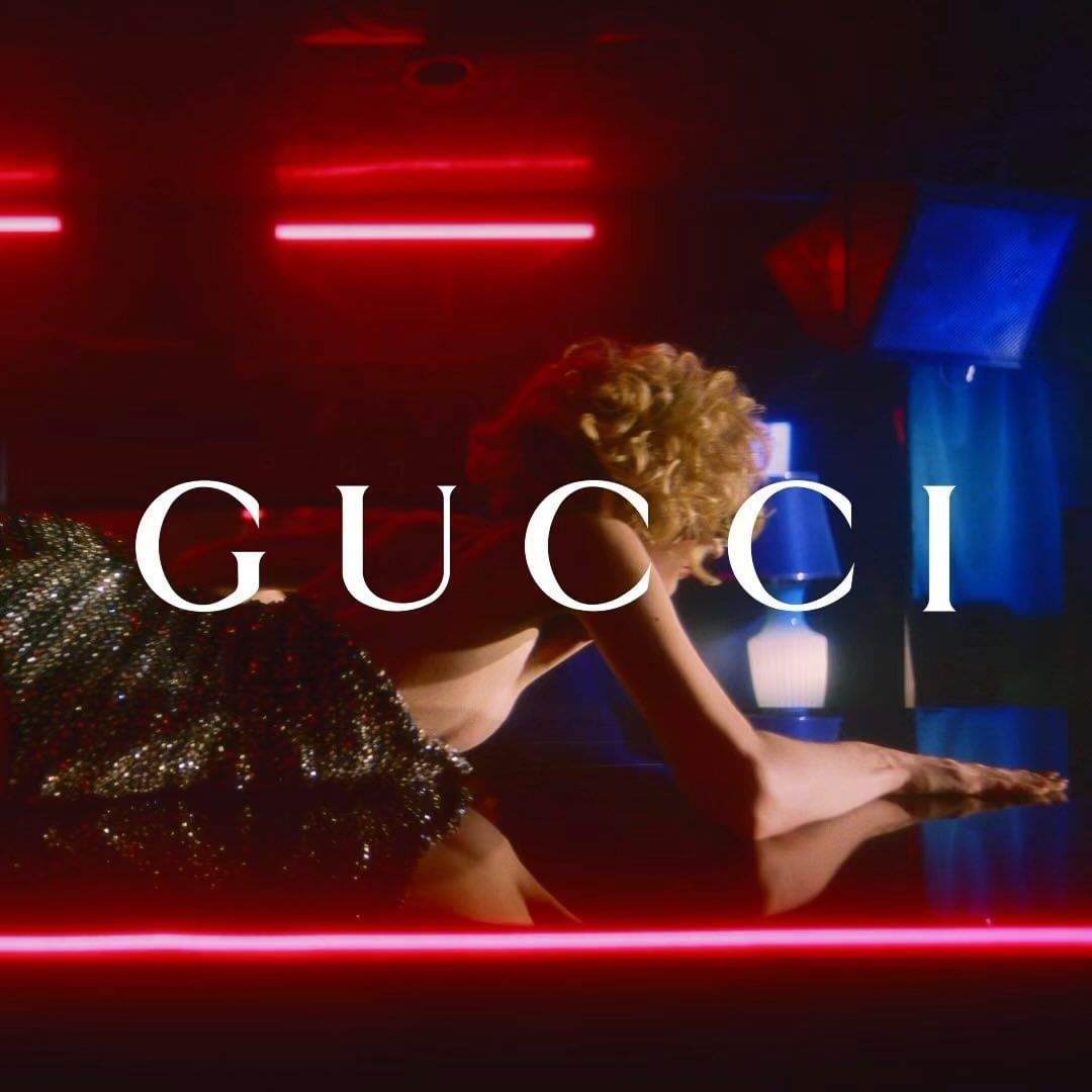 Adult female model in a glittering dress poses low to the ground in a dark photo studio with red and blue neon lighting and Gucci logo.