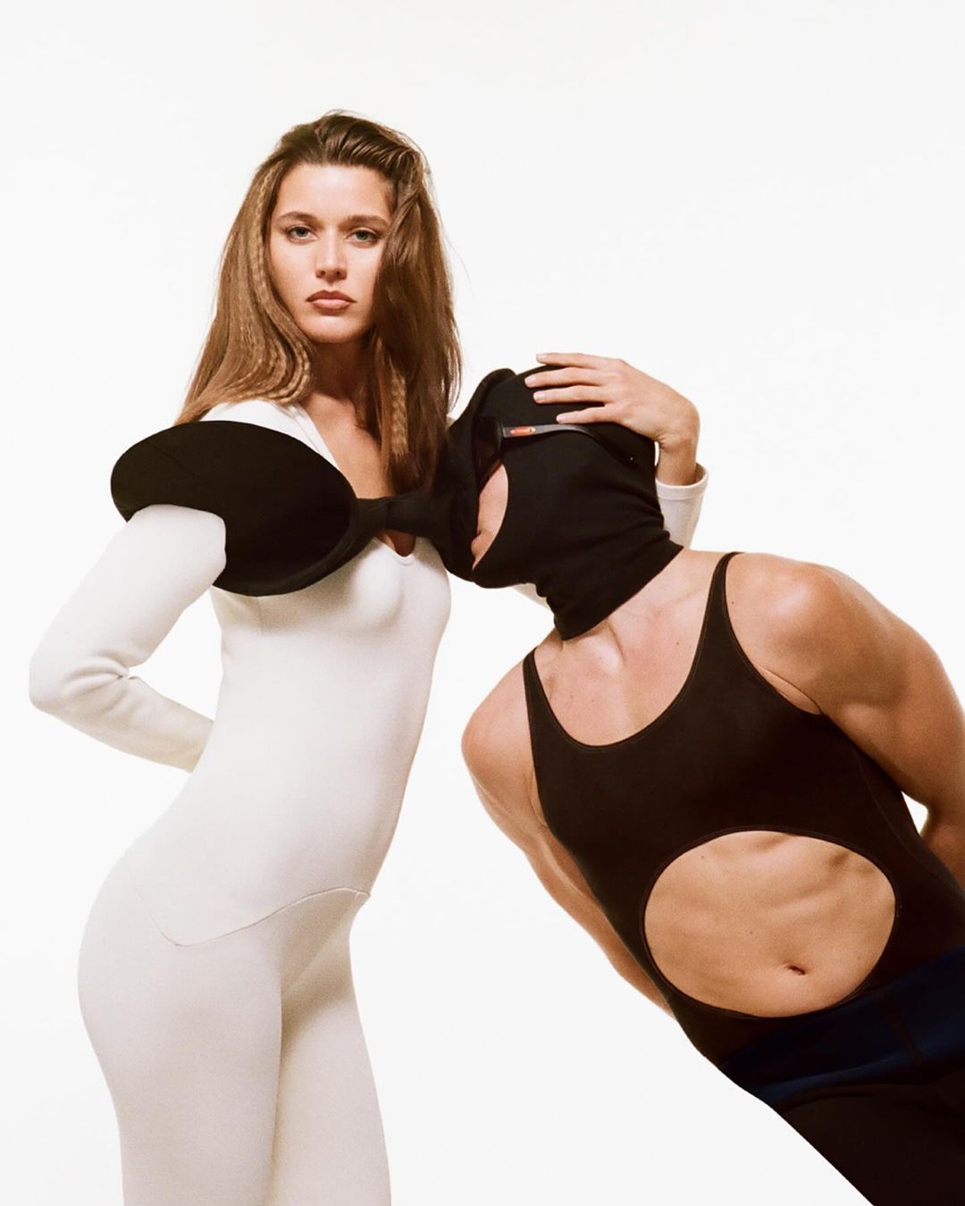 Two people in studio wearing black and white form fitting outfits. One person is wearing a face covering in a studio setting.