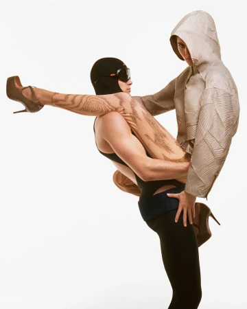 Two fashion models pose in a studio with creative costuming; a man carries a woman with a patterned leg covering. The woman wears a hooded top.