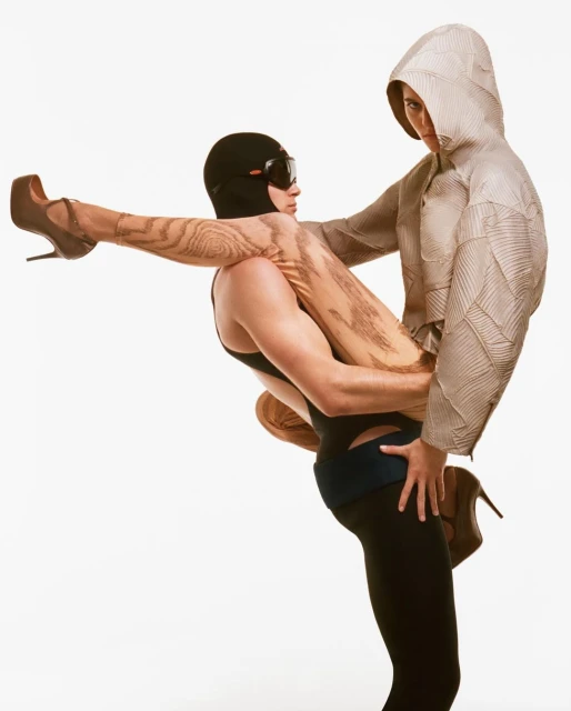 Fashion shot featuring two models, one carrying the other. The carrier wears a black balaclava, while the other has a grey geometric pattern hood.