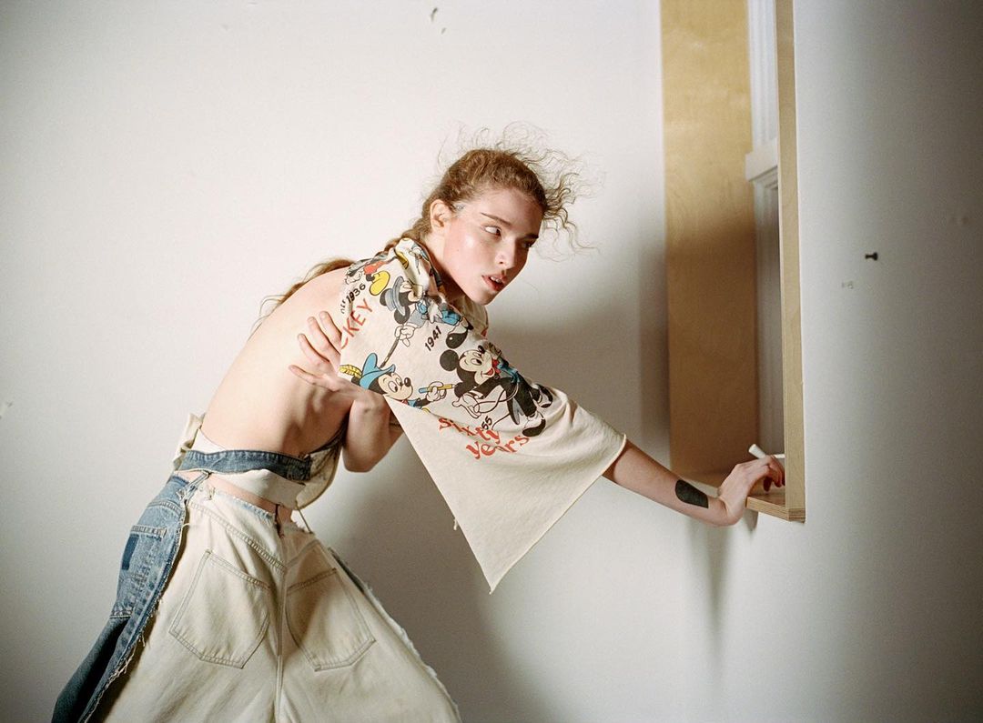 Woman in a Mickey Mouse shirt and disassembled jeans poses in a room with white walls and a light wood window frame.