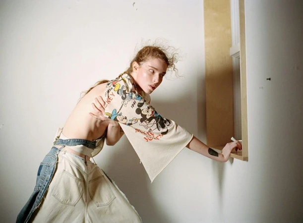 Woman in a Mickey Mouse shirt and disassembled jeans poses in a room with white walls and a light wood window frame.