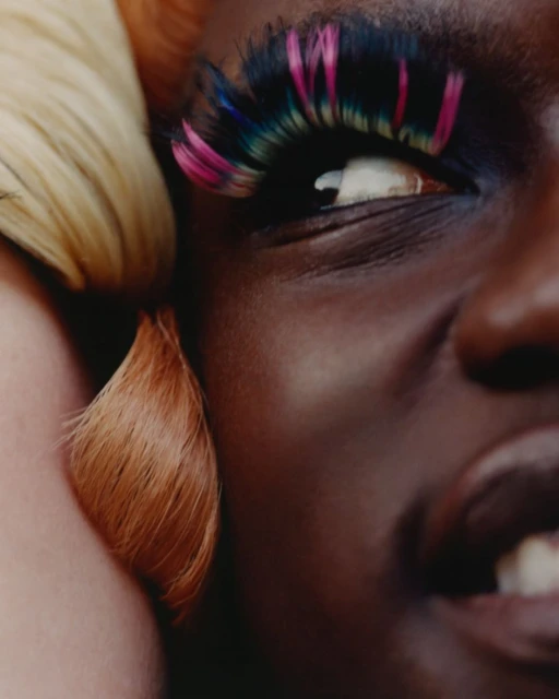 Close-up beauty shot of a smiling Black female model wearing large, colorful false eyelashes, with platinum and orange-tinted hair.