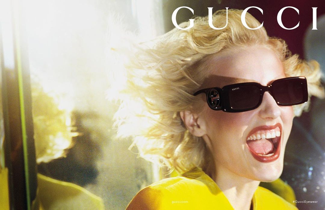 Blonde woman in yellow jacket and dark sunglasses with visible brand logo smiles in bright outdoor fashion shot for Gucci Eyewear.