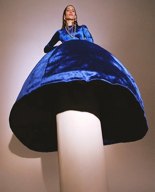 Eye-level portrait of a woman in a blue velvet dress posing on a mushroom-shaped stool against a neutral backdrop in a studio setting.