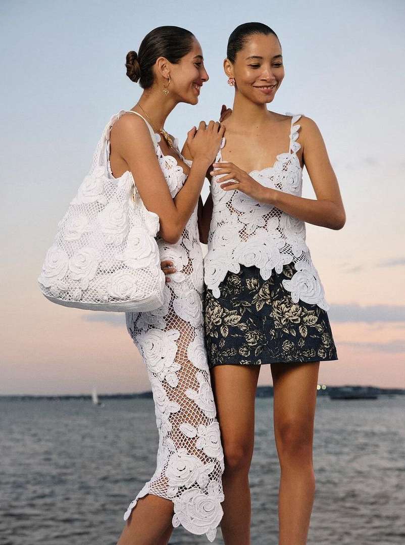 Two smiling women pose outdoors wearing white crochet dresses. One carries a matching tote bag. Ocean and sky in the background.
