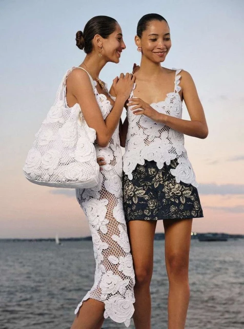 Two smiling women pose outdoors wearing white crochet dresses. One carries a matching tote bag. Ocean and sky in the background.