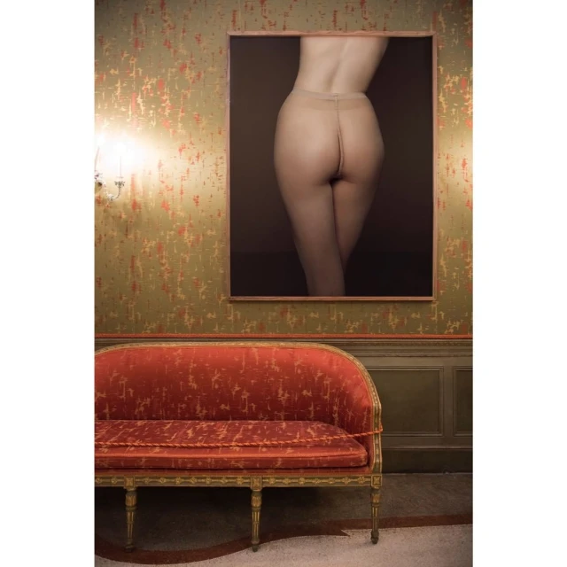 Photograph of an antique-style red sofa, gold ornate details, wallpaper with a nude photograph in a frame, and sconce wall lighting.