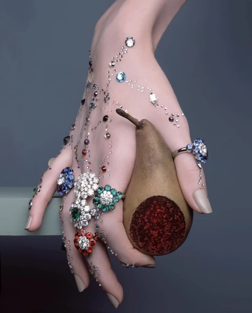 Close-up of a hand adorned with jeweled rings and small gems, delicately holding a pear against a grey backdrop, showcasing jewelry.