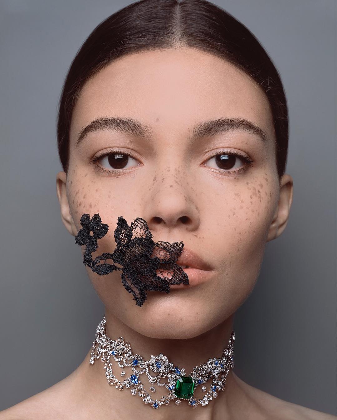 Close-up portrait of a young woman with freckles wearing a jeweled necklace, and black lace floral embellishment on her face, looking forward.
