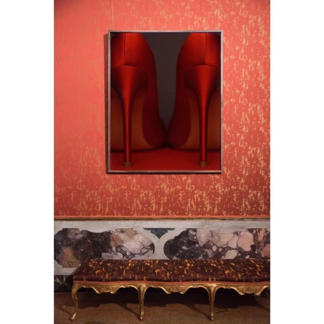 Framed art print of two red high-heel shoes hanging on a red patterned wall above a gold-legged bench with a brown patterned cushion.