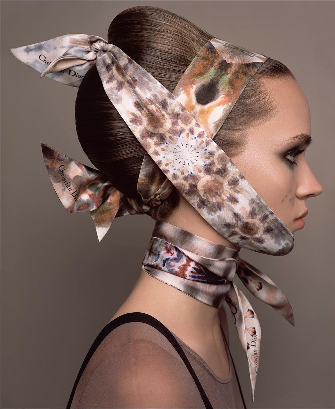 Fashion shot of a woman with dark hair and dramatic eye makeup wearing patterned headscarf and sheer top against a grey background.