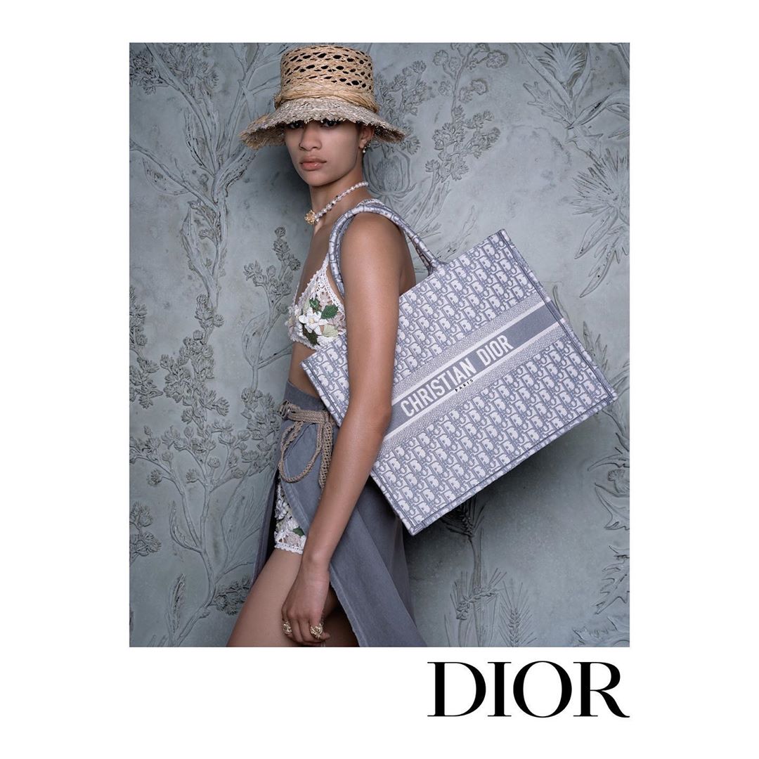 A tanned woman in a summer-style ensemble holds a branded tote bag against a floral backdrop, evoking high-fashion and vacation chic.