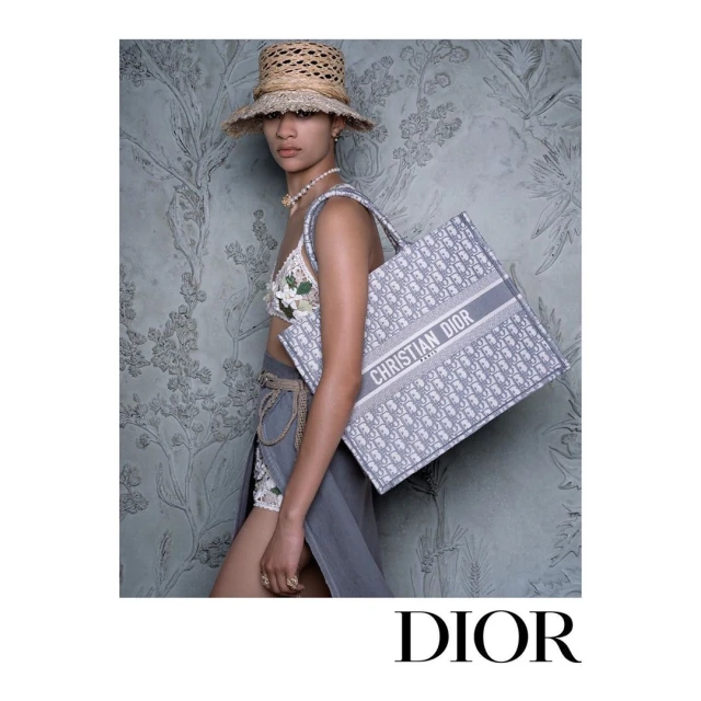 A tanned woman in a summer-style ensemble holds a branded tote bag against a floral backdrop, evoking high-fashion and vacation chic.